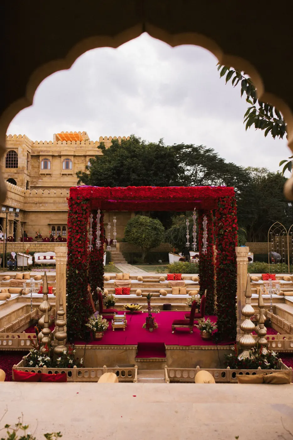 A red floral mandap in an ornate courtyard, framed by a carved arch
